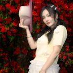 Fashionable woman in a garden holding a hat, surrounded by vivid red flowers.